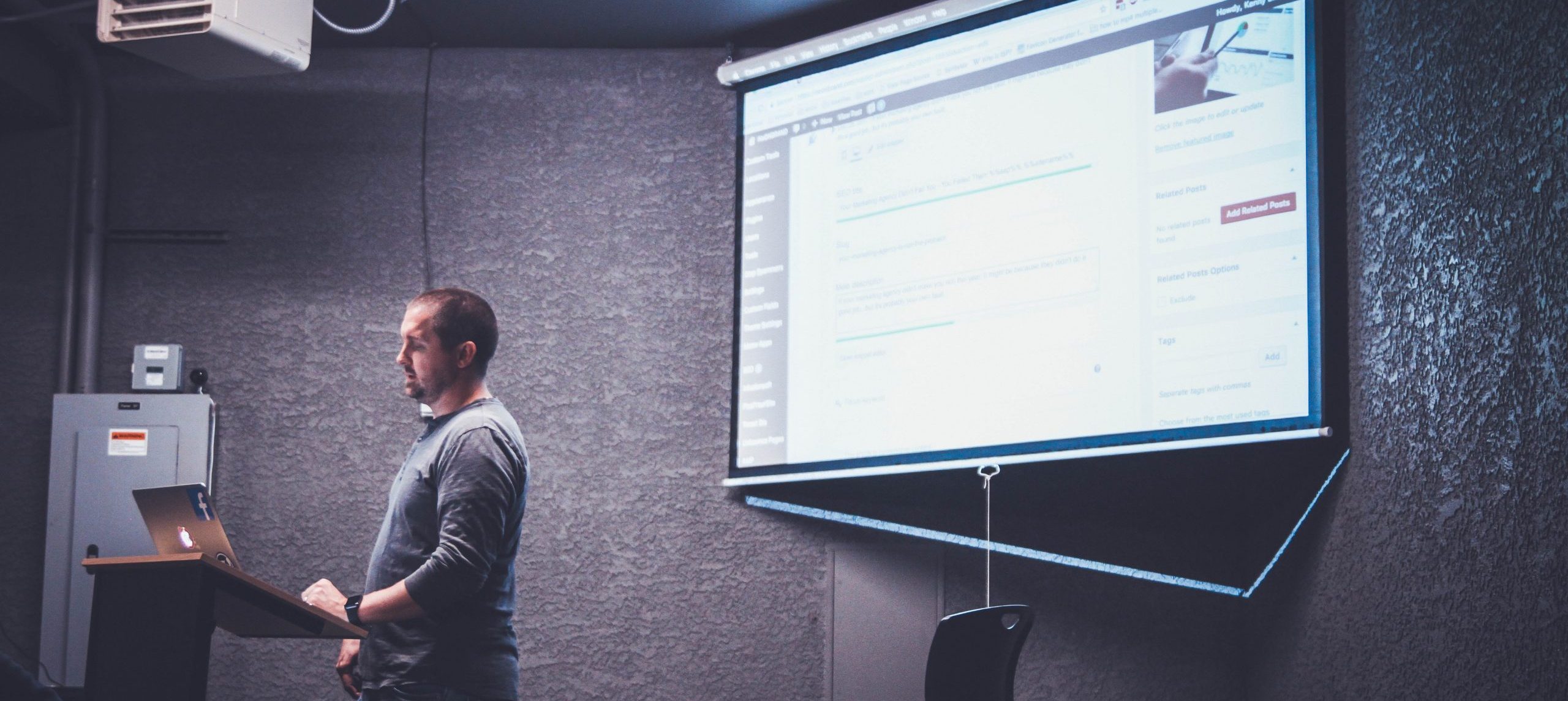 Ein Mann, der links in einem Raum vor einem Pult mit einem Laptop steht, rechts eine PowerPoint-Folie auf einer Projektorleinwand.