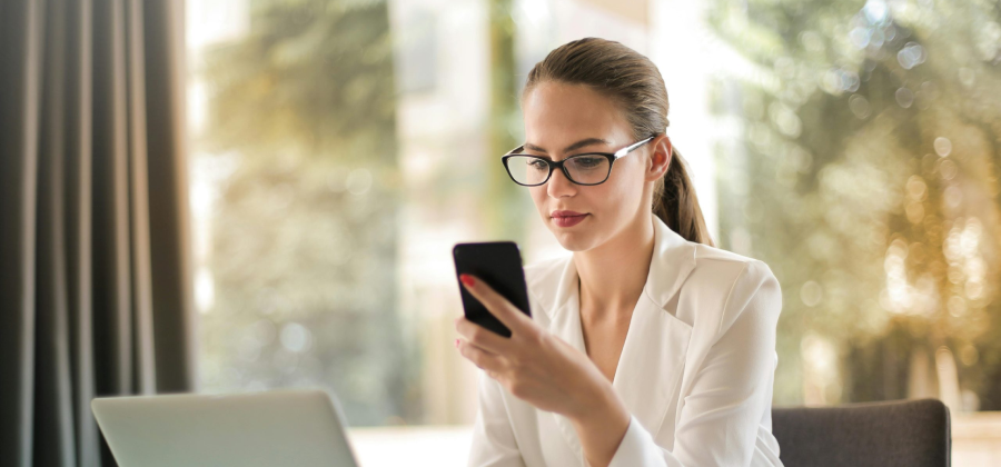 A woman in a white blazer, wearing glasses, is looking at her smartphone and sitting at a desk with a laptop, possibly researching how to make online courses more interactive.