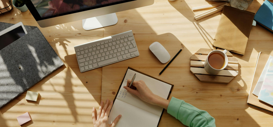 Person jotting ideas in a notebook on a wooden desk, accompanied by a computer, keyboard, coffee, and books. As sunlight casts shadows across the surface, inspiration strikes on how to make online courses more interactive.