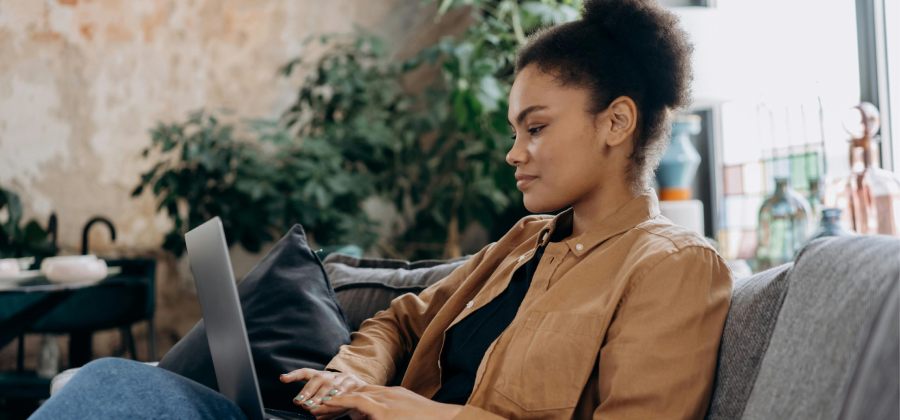 A woman sits on a couch surrounded by lush indoor plants, using her laptop to explore ways to make online courses more interactive.