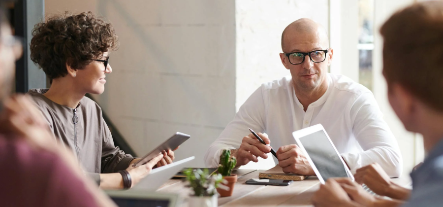 A group of people sitting at a table with electronic devices and notebooks, deep in discussion about the new corporate training platform. The person in the center is holding a pen.