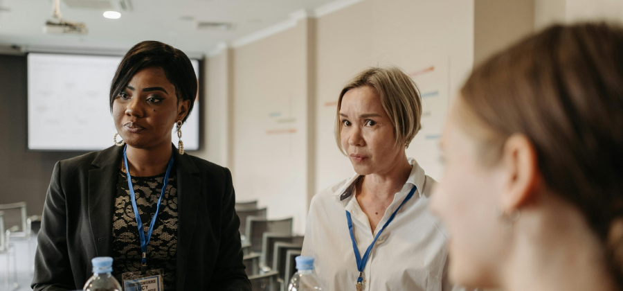 Three individuals wearing conference badges engage in a lively discussion on how to make online courses more interactive, as they stand in a meeting room with a presentation screen in the background.