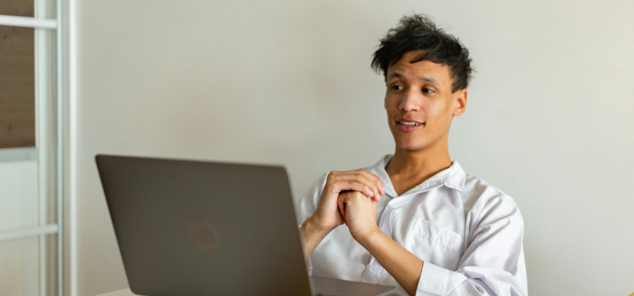 A person in a white shirt sits at a table, hands clasped, examining the laptop screen with a neutral expression, pondering how to make online courses more interactive.