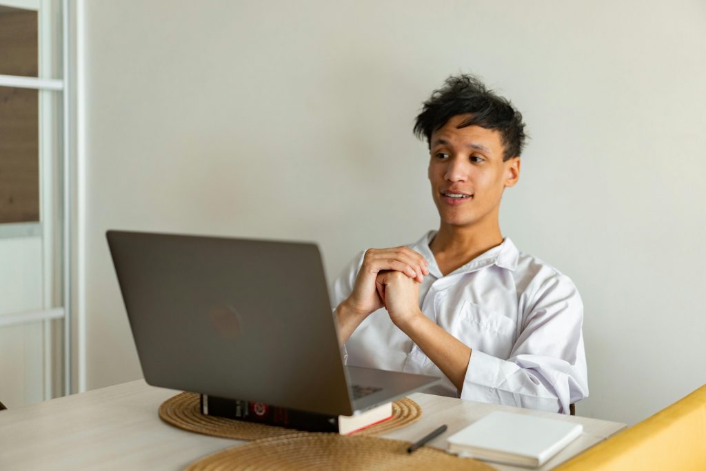 A person with tousled hair sits at a table, intently looking at a laptop. Wearing a white shirt, they have a notebook and pen nearby as they delve into e-learning localization tasks.