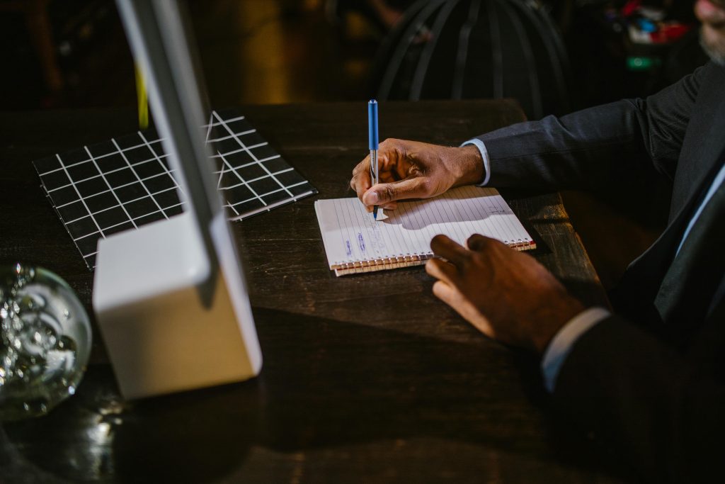 A person is thoughtfully writing in a notebook with a pen at their desk, surrounded by an array of digital tools for e-learning localization, including a monitor and grid notebook.