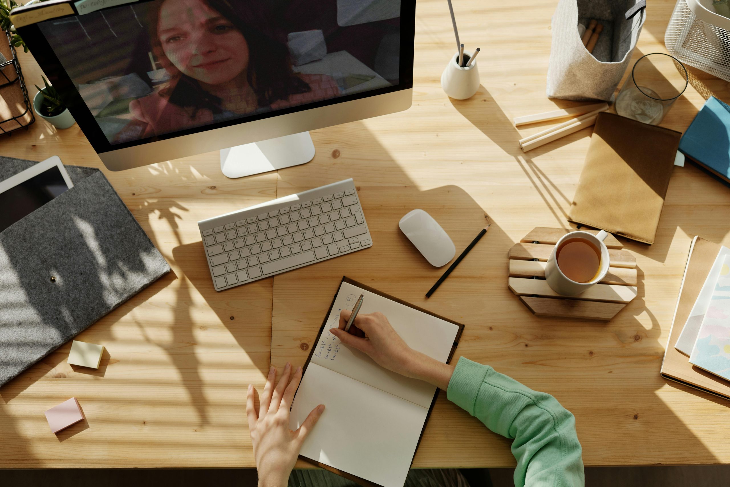 A person writes in a notebook while participating in a video call on a computer at a wooden desk with a keyboard, mouse, coffee, sticky notes, and stationery items.