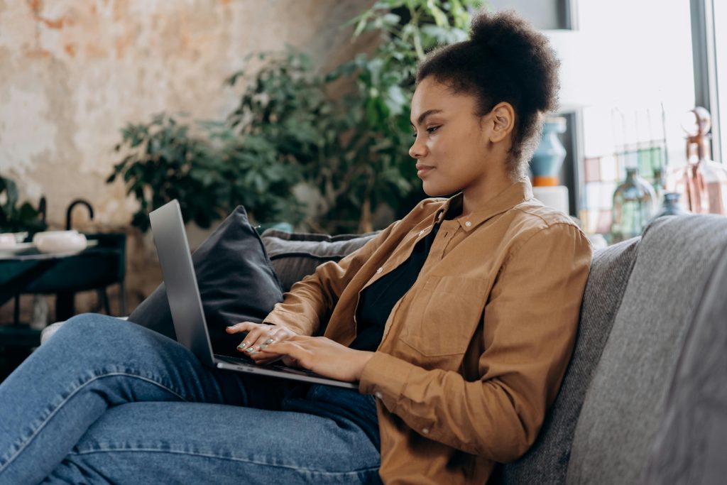 A person sits on a couch, engaged with their laptop on a corporate training platform. Surrounded by lush plants, they wear a brown shirt and blue jeans, blending comfort with productivity.