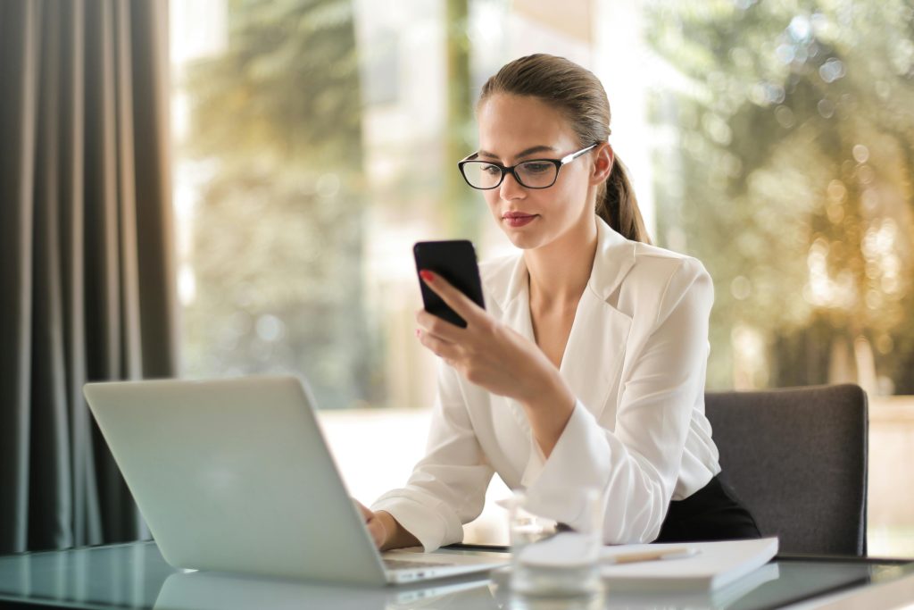 A woman in a white blouse sits at a desk, creating training materials. She glances at her smartphone, with an open laptop and a glass of water nearby.