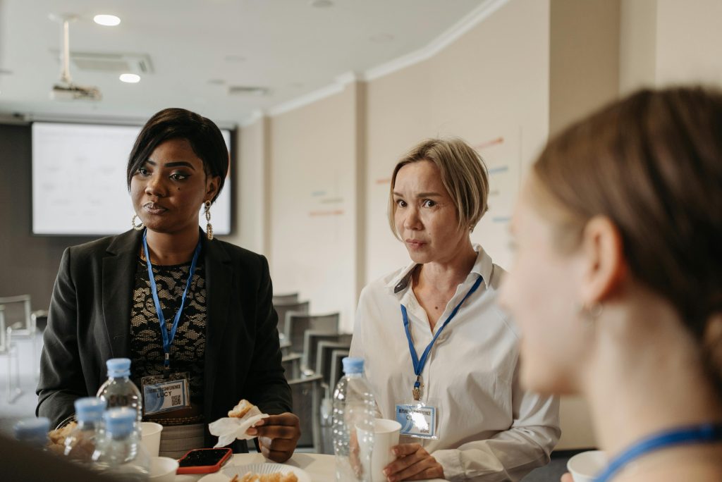 Three women with name tags stand in a room near a round table filled with water bottles, paper cups, and snacks, appearing to engage in conversation. They seem to be discussing the latest corporate training models for improving workplace productivity.