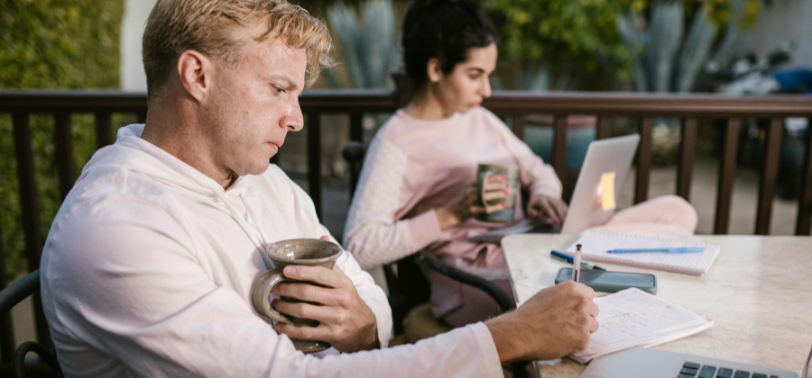 Two individuals sit on a patio, engrossed in their tasks. One writes in a notebook with a mug in hand, while the other types on a laptop. Their focus suggests an adaptive learning design approach, seamlessly blending work and creativity in this serene setting.